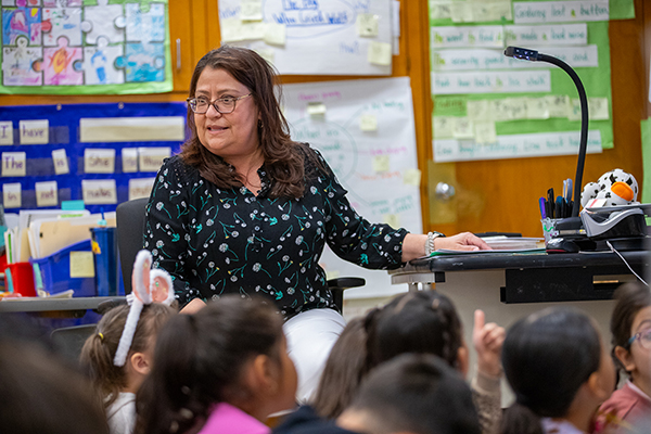 A woman with brown hair and glasses talks to young students sitting on the floor